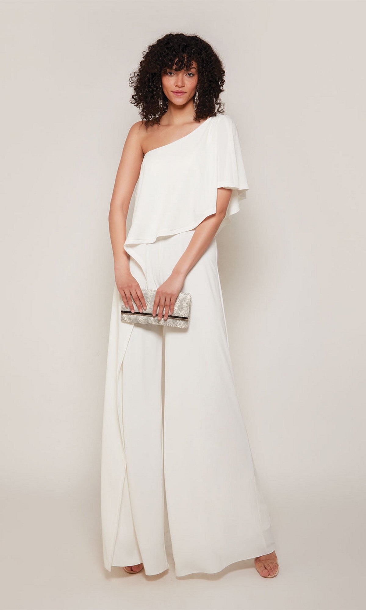 A woman stands against a light background wearing the Alyce 70049 long formal jumpsuit in ivory and holding a silver clutch bag.